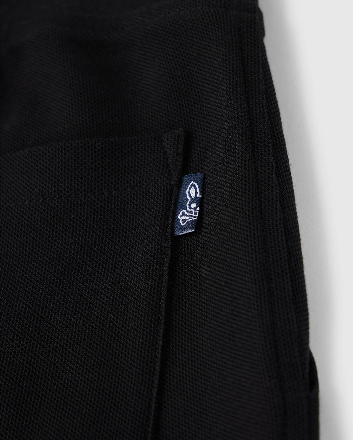 Close-up of a black KIDS STANFORD LIGHT PIQUE SWEATSHORT - B0R344B200 with a small pocket. A blue tag with a white rabbit skull and crossbones logo is attached to the pocket's edge. Made from Pima cotton piqué, this stylish wardrobe staple by Psycho Bunny is set against a plain white background.