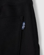 Close-up of a black KIDS STANFORD LIGHT PIQUE SWEATSHORT - B0R344B200 with a small pocket. A blue tag with a white rabbit skull and crossbones logo is attached to the pocket's edge. Made from Pima cotton piqué, this stylish wardrobe staple by Psycho Bunny is set against a plain white background.