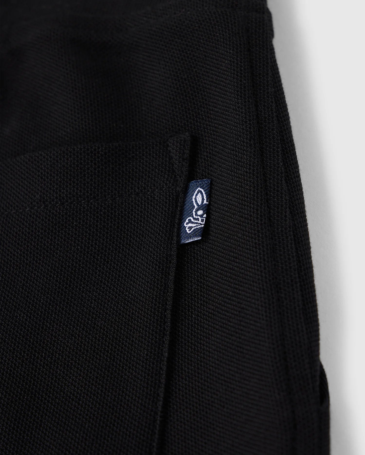Close-up of a black KIDS STANFORD LIGHT PIQUE SWEATSHORT - B0R344B200 with a small pocket. A blue tag with a white rabbit skull and crossbones logo is attached to the pocket's edge. Made from Pima cotton piqué, this stylish wardrobe staple by Psycho Bunny is set against a plain white background.