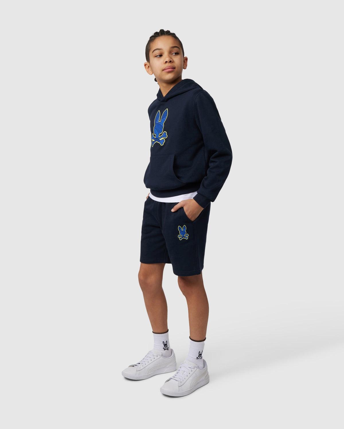 A young person stands confidently in a navy hoodie and Psycho Bunny's KIDS LENOX SWEATSHORT - B0R377B200, featuring naturally-breathable fleece with a colorful cartoon graphic. Their hands rest in the pockets, complemented by white sneakers and matching socks, ideal for an active lifestyle on a light gray backdrop.