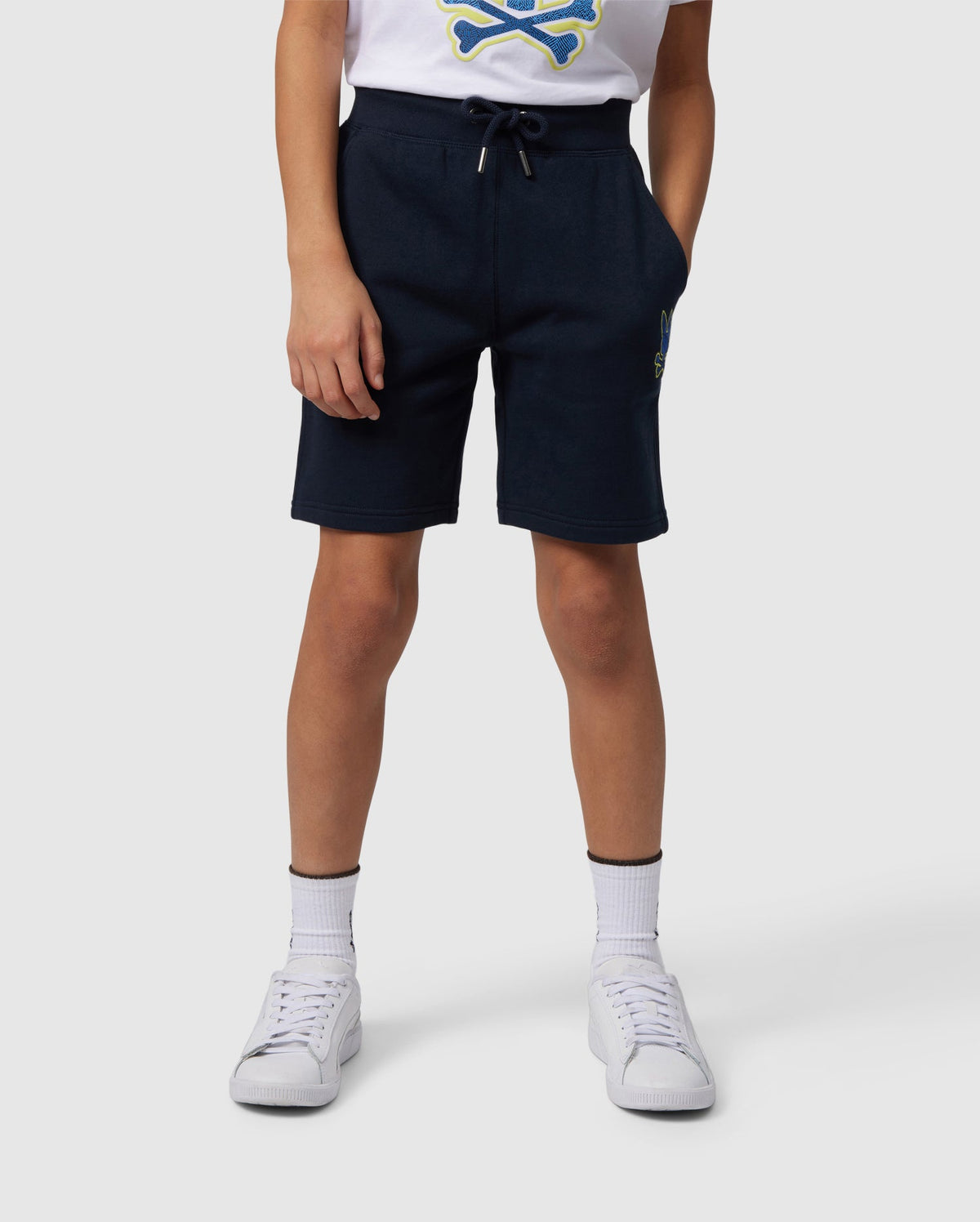 Dressed in a white t-shirt featuring a skull and crossbones, navy blue Psycho Bunny Kids Lenox Sweatshorts B0R377B200, white socks, and sneakers against a plain gray backdrop. The naturally breathable fleece and timeless style complement an active lifestyle.