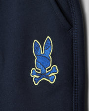 Close-up of Psycho Bunny's navy kids' Lenox sweatshort (B0R377B200) showing a blue bunny head patch with yellow outline and crossed bones near the pocket seam. Made from breathable fleece, it's ideal for an active lifestyle.