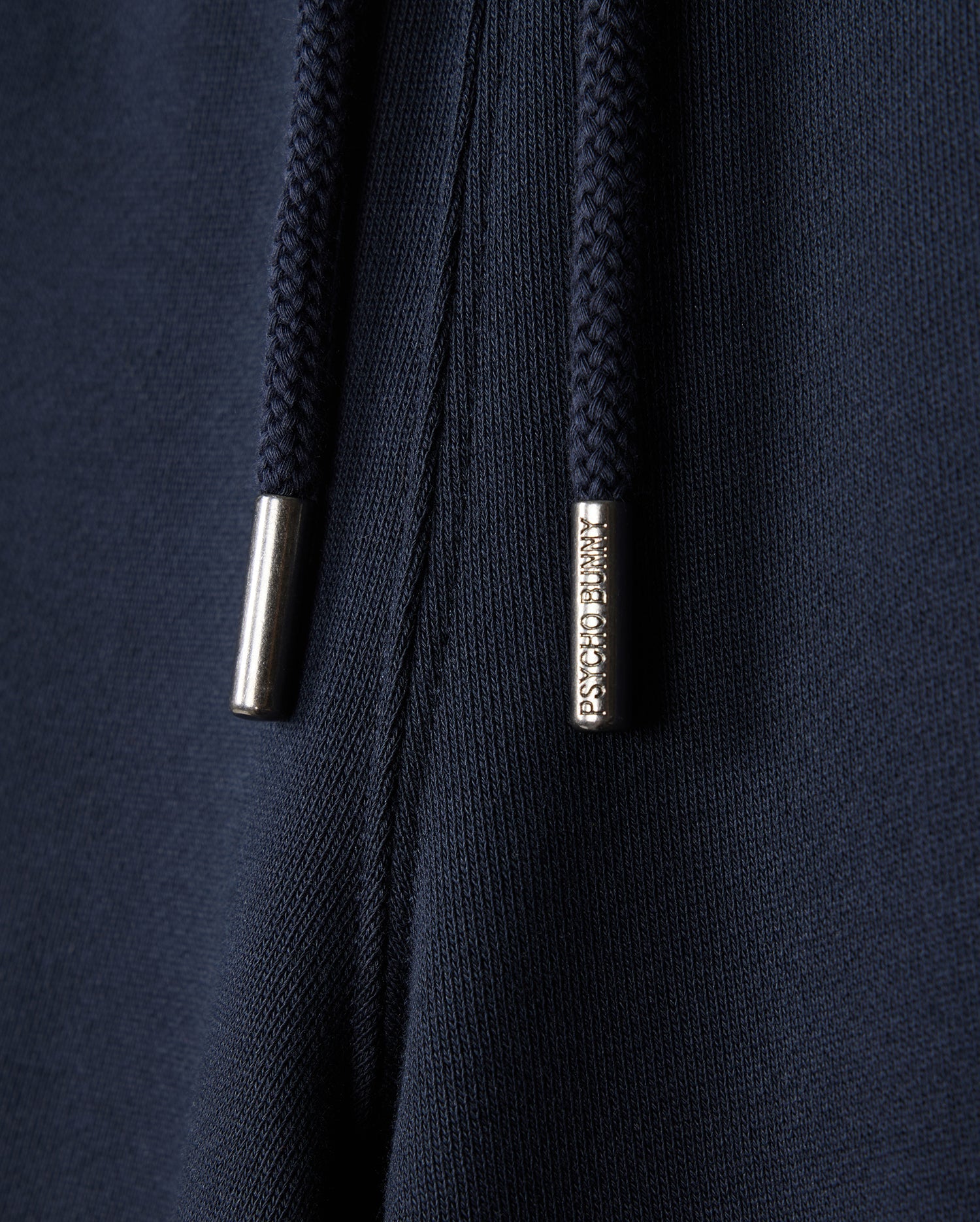 Close-up of dark blue fabric sweatshorts with navy drawstrings featuring "Psycho Bunny" engraved metal tips. This naturally breathable fleece from Psycho Bunny's KIDS LENOX SWEATSHORT - B0R377B200 collection is perfect for an active lifestyle.