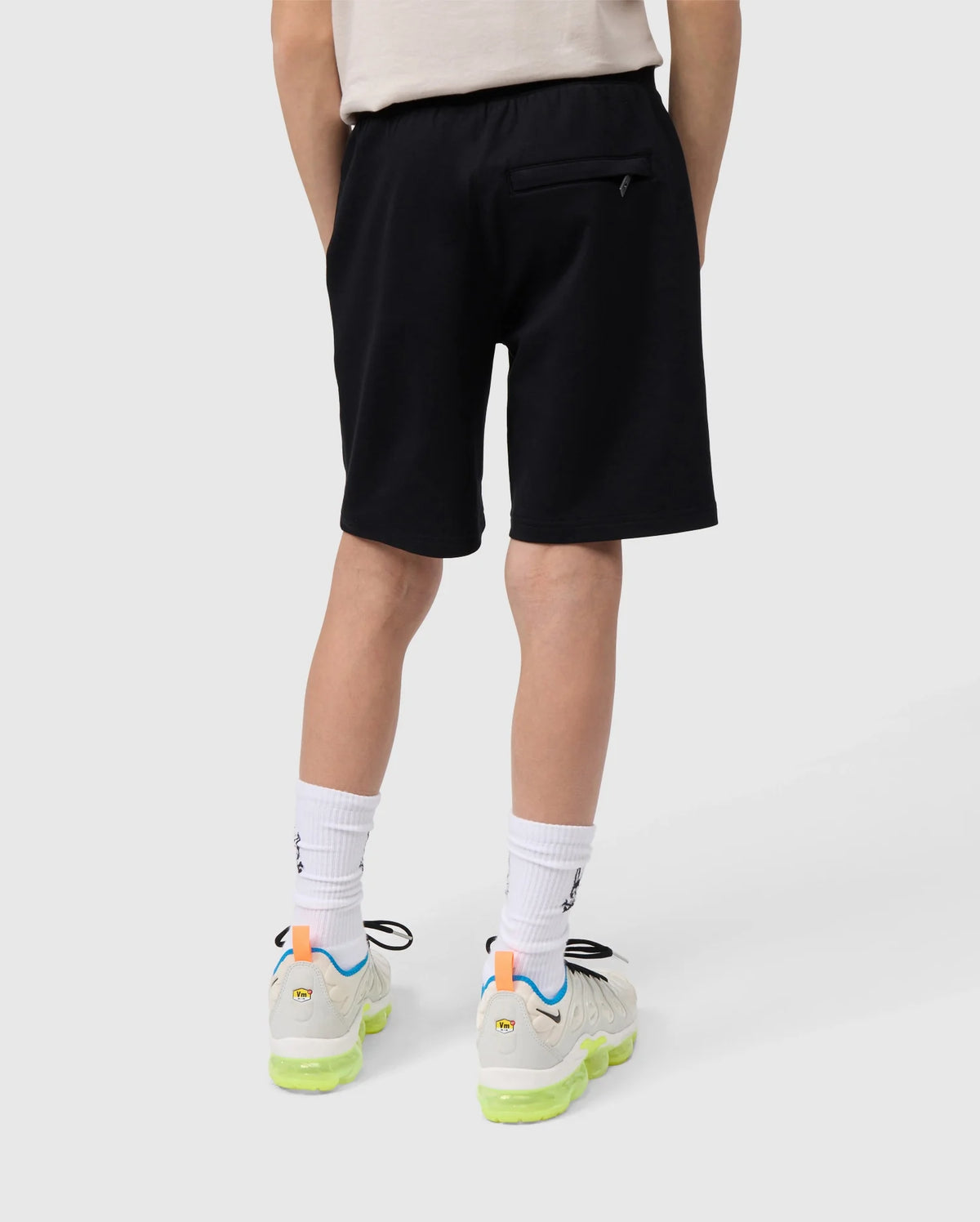 A child is standing against a plain background, facing away from the camera. They are wearing a white T-shirt and comfortable black KIDS LIVINGSTON TERRY SWEATSHORT - B0R408B200 by Psycho Bunny with a zippered back pocket. Also styled in white socks and white sneakers with neon yellow-green accents on the soles, they seem ready for play in Micro French Terry.