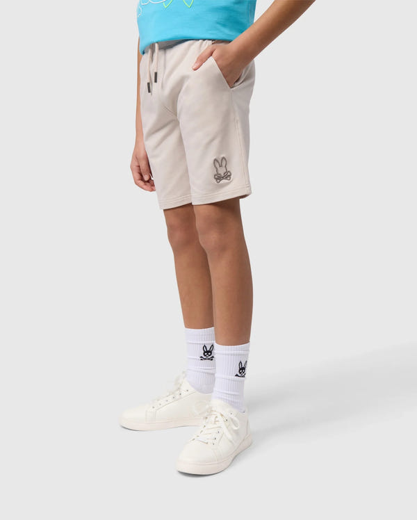 A person standing wearing beige Psycho Bunny Livingston terry sweatshorts with a small bunny logo, white socks with a logo at the top, and white sneakers. Only the lower half of the body is visible.