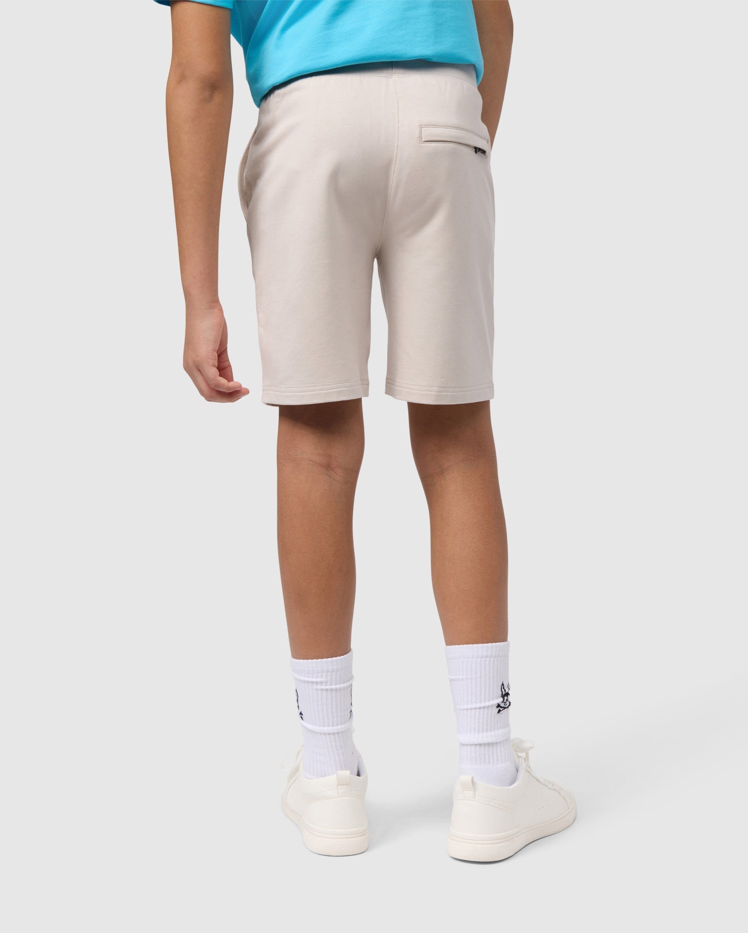 A person wearing Psycho Bunny KIDS LIVINGSTON TERRY SWEATSHORT - B0R408B200, white socks with a black emblem, and white sneakers, viewed from behind from the waist down.