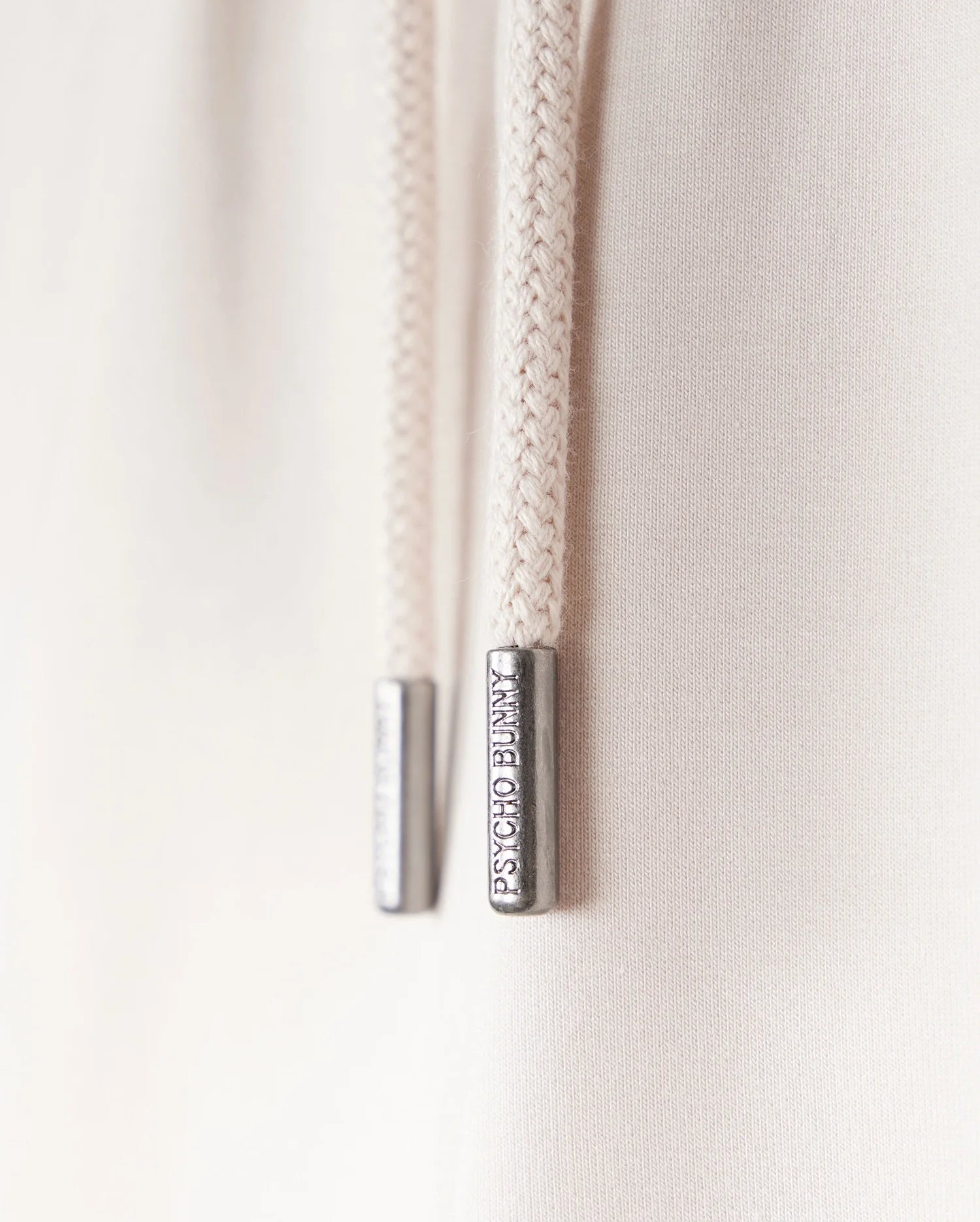 Close-up of two cream-colored drawstring cords with metal tips engraved with "LIMITED EDITION." The cords, part of a Psycho Bunny KIDS LIVINGSTON TERRY SWEATSHORT - B0R408B200, hang against a soft, light background.