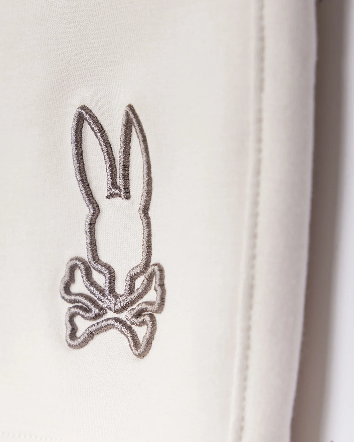 A close-up of a grey embroidered logo depicting a stylized rabbit with long ears and a bow tie, stitched onto a white Psycho Bunny Livingston Terry Sweatshort background.