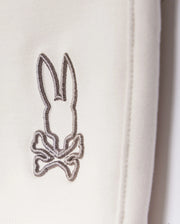 A close-up of a grey embroidered logo depicting a stylized rabbit with long ears and a bow tie, stitched onto a white Psycho Bunny Livingston Terry Sweatshort background.