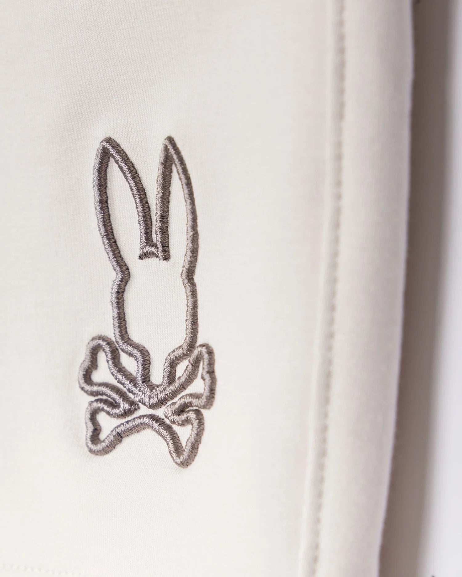 A close-up of a grey embroidered logo depicting a stylized rabbit with long ears and a bow tie, stitched onto a white Psycho Bunny Livingston Terry Sweatshort background.