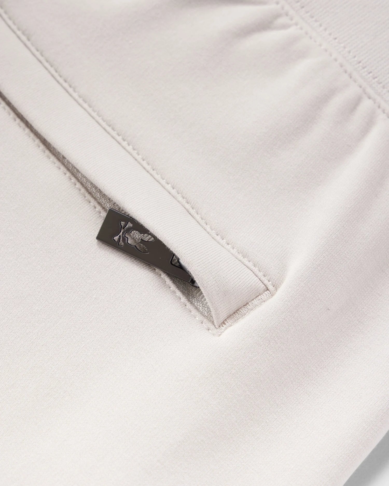 Close-up view of a beige micro French terry fabric with a metal zipper partially unzipped, showing a detailed floral pattern on a small fabric tag from Psycho Bunny's KIDS LIVINGSTON TERRY SWEATSHORT - B0R408B200.