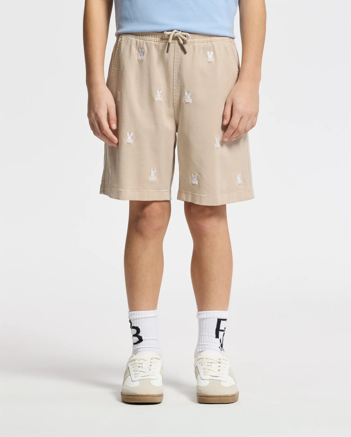 A person wears Psycho Bunny beige KIDS ALL OVER WILLIS STRETCH TENCEL SHORTS with a bunny pattern, hands in pockets; paired with a light blue shirt, white socks featuring black text, and white sneakers, against a plain white background.