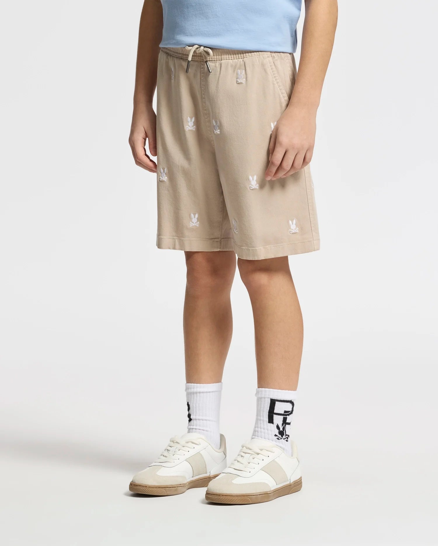 A person sports the KIDS ALL OVER WILLIS STRETCH TENCEL SHORT by Psycho Bunny, a light blue shirt, white socks with black designs, and white sneakers with beige accents. Beige shorts feature an elastic waist and small white bunny patterns. Only their midsection and legs are visible against a plain background.