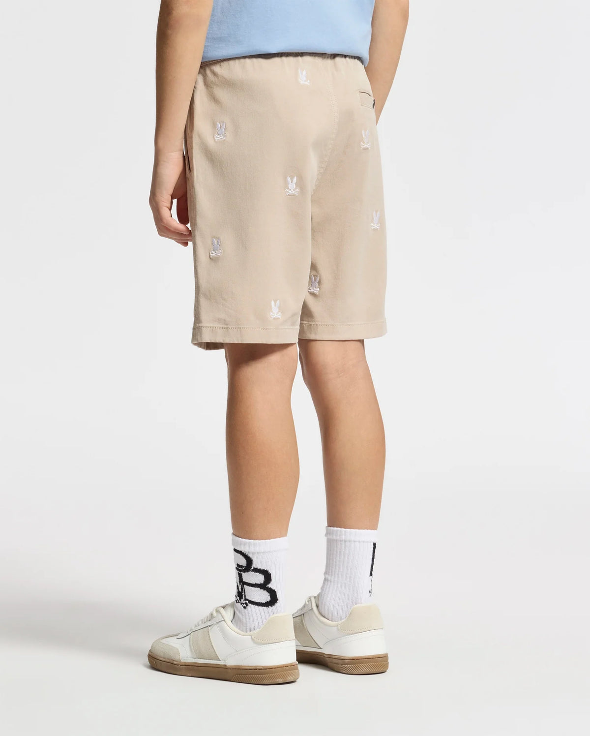 Wearing Psycho Bunny's KIDS ALL OVER WILLIS STRETCH TENCEL SHORT in beige with a bunny pattern, paired with white socks adorned with black letters and crisp white sneakers, they stand against a pristine white backdrop facing away.