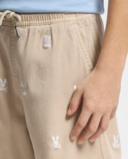 A person wears Psycho Bunny's KIDS ALL OVER WILLIS STRETCH TENCEL SHORTS, showcasing embroidered bunny and skull motifs on tan fabric. These regular fit shorts feature a stretch Tencel blend, elastic waistband with drawstring, and are paired with a glimpse of a light blue top.