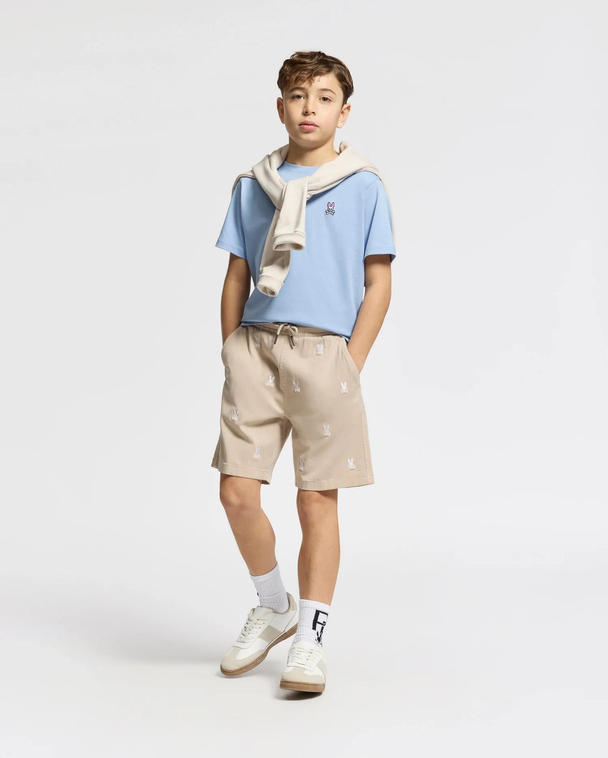 A boy poses against a plain background, wearing a light blue T-shirt with a white sweater draped over his shoulders. He sports tan Psycho Bunny KIDS ALL OVER WILLIS STRETCH TENCEL SHORTS featuring an elastic waist and subtle bunny pattern, paired with white socks and sneakers, hands casually in pockets.
