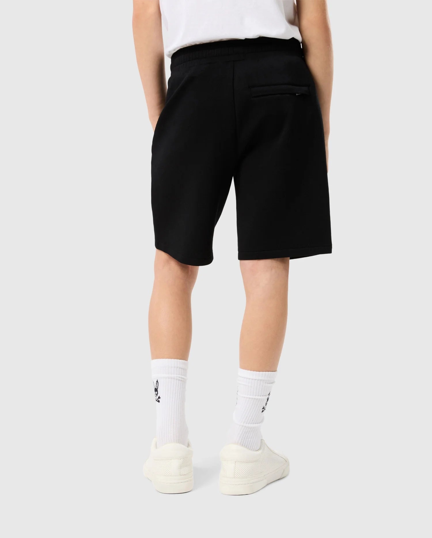 Rear view of a person wearing Psycho Bunny KIDS LYONS SWEATSHORT - B0R578C200, white socks with a logo near the top, and cream-colored sneakers, standing against a light background.