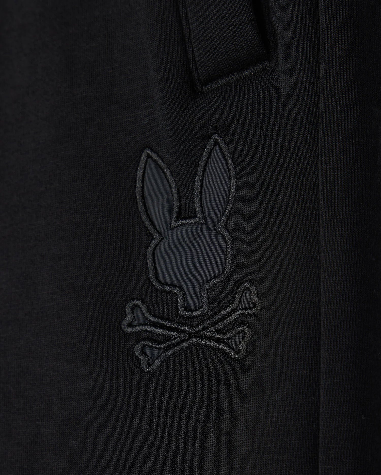 Close-up of a black cotton blend Kids Lyons Sweatshort by Psycho Bunny with an embroidered patch featuring a stylized skull and crossbones design shaped like a rabbit head.