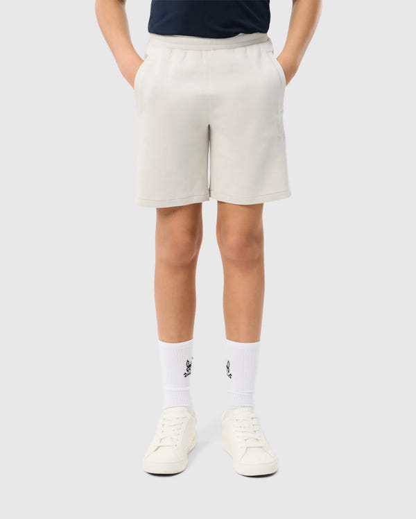 Person wearing beige Psycho Bunny KIDS LYONS SWEATSHORT - B0R578C200, a black t-shirt, white socks with a small black logo, and white sneakers. The person is standing with hands in pockets against a plain white background.