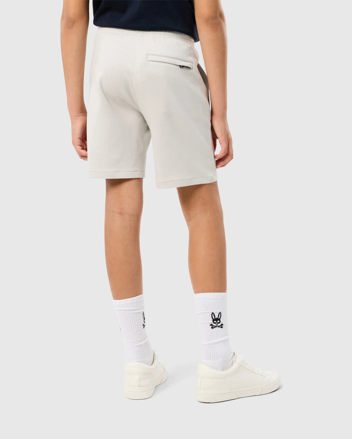 A person is standing, showcasing the back view of their outfit. They are wearing light-colored Psycho Bunny KIDS LYONS SWEATSHORT - B0R578C200, white socks with black logos, and white sneakers. The background is a plain, light grey.