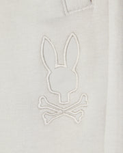 Close-up of a white Kids casual short made from soft stretch modal-cotton-blend jersey, featuring a minimalist embroidered design resembling a rabbit head with crossed bones underneath. The white stitching on the KIDS LYONS SWEATSHORT - B0R578C200 by Psycho Bunny creates a subtle, monochromatic effect.
