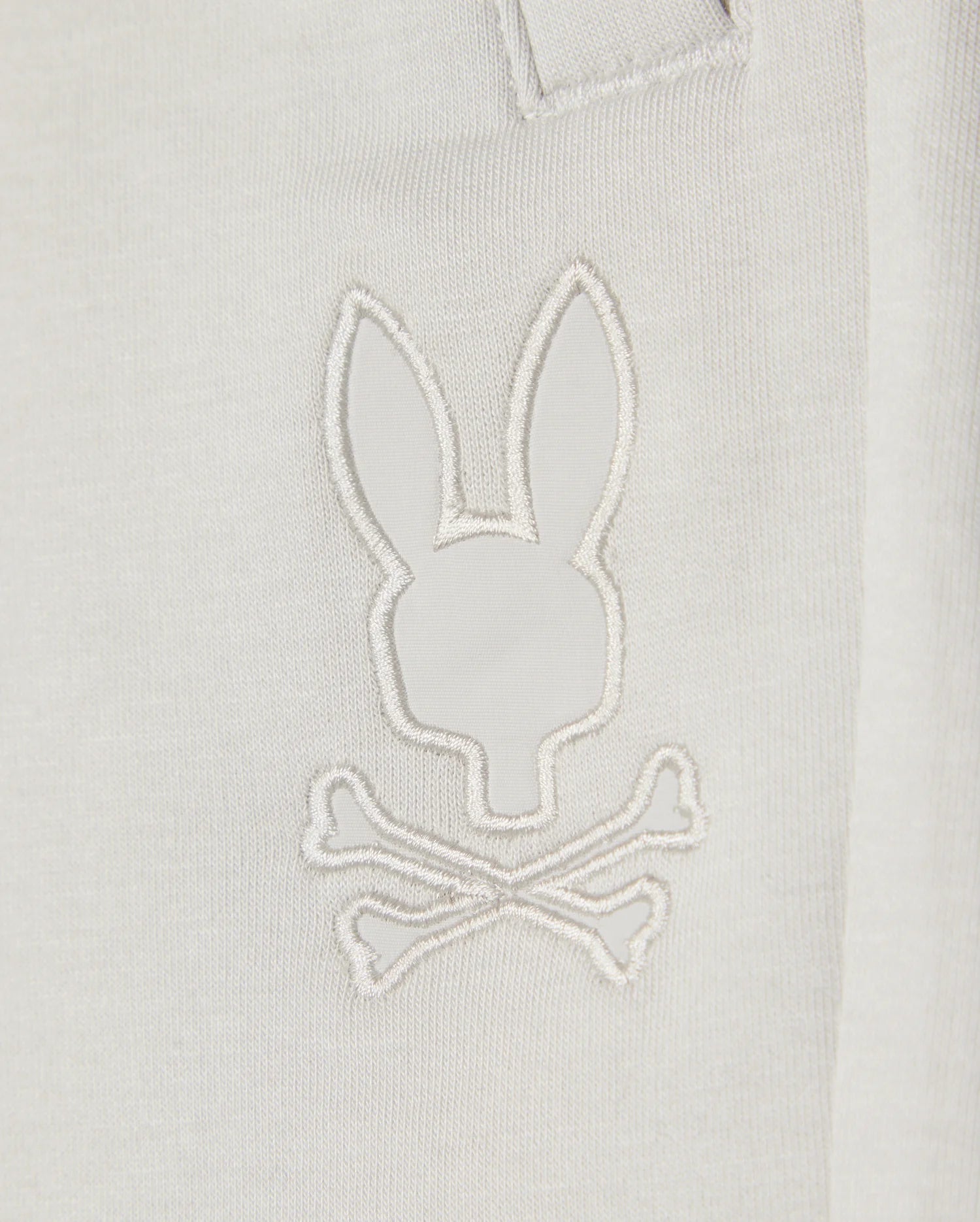 Close-up of a white Kids casual short made from soft stretch modal-cotton-blend jersey, featuring a minimalist embroidered design resembling a rabbit head with crossed bones underneath. The white stitching on the KIDS LYONS SWEATSHORT - B0R578C200 by Psycho Bunny creates a subtle, monochromatic effect.