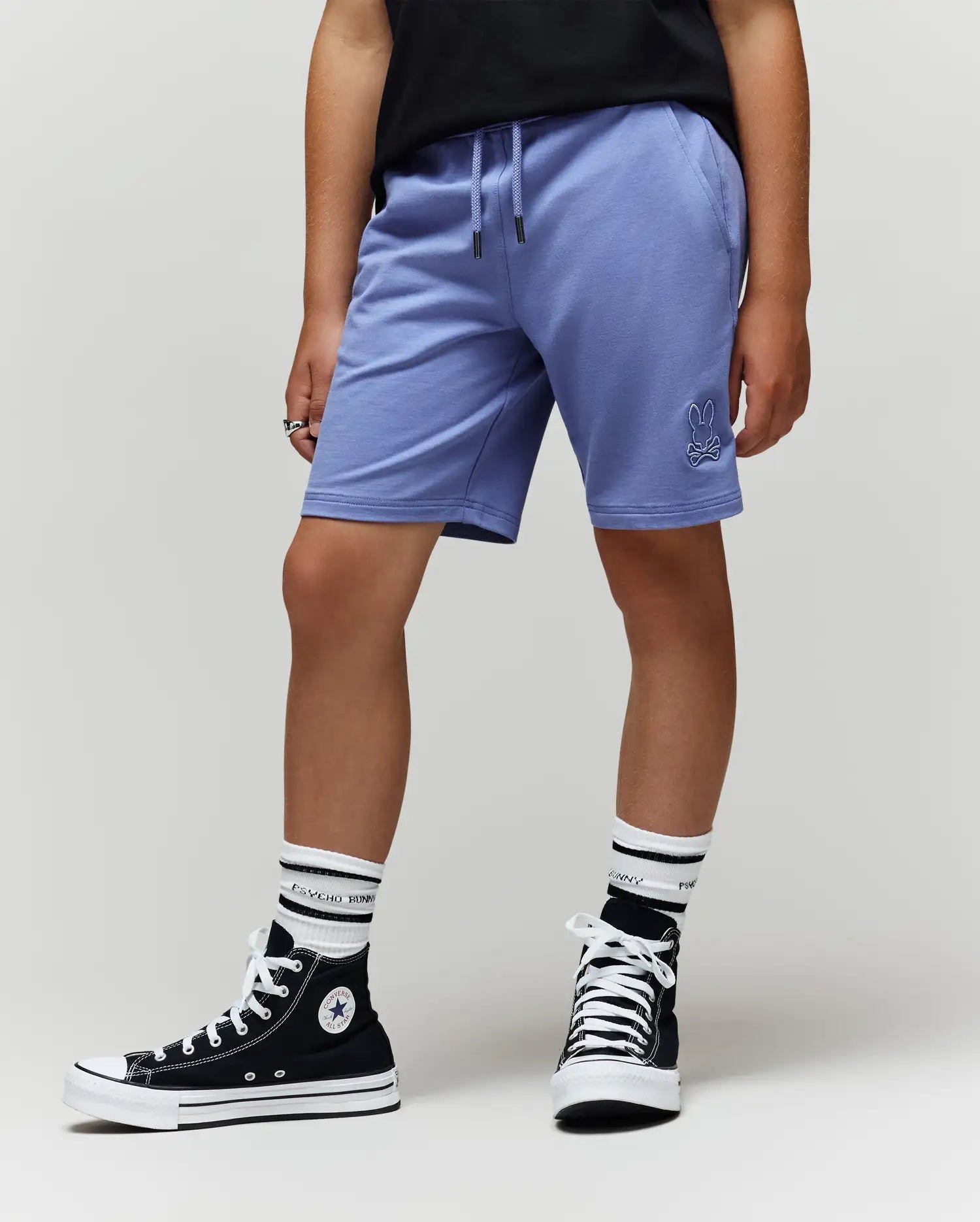 A person wears lavender Psycho Bunny KIDS LEVANZO FRENCH TERRY SWEATSHORTS (B0R578F200) with an embroidered Bunny, a black t-shirt, white socks with black stripes, and black high-top Converse sneakers against a plain light background.