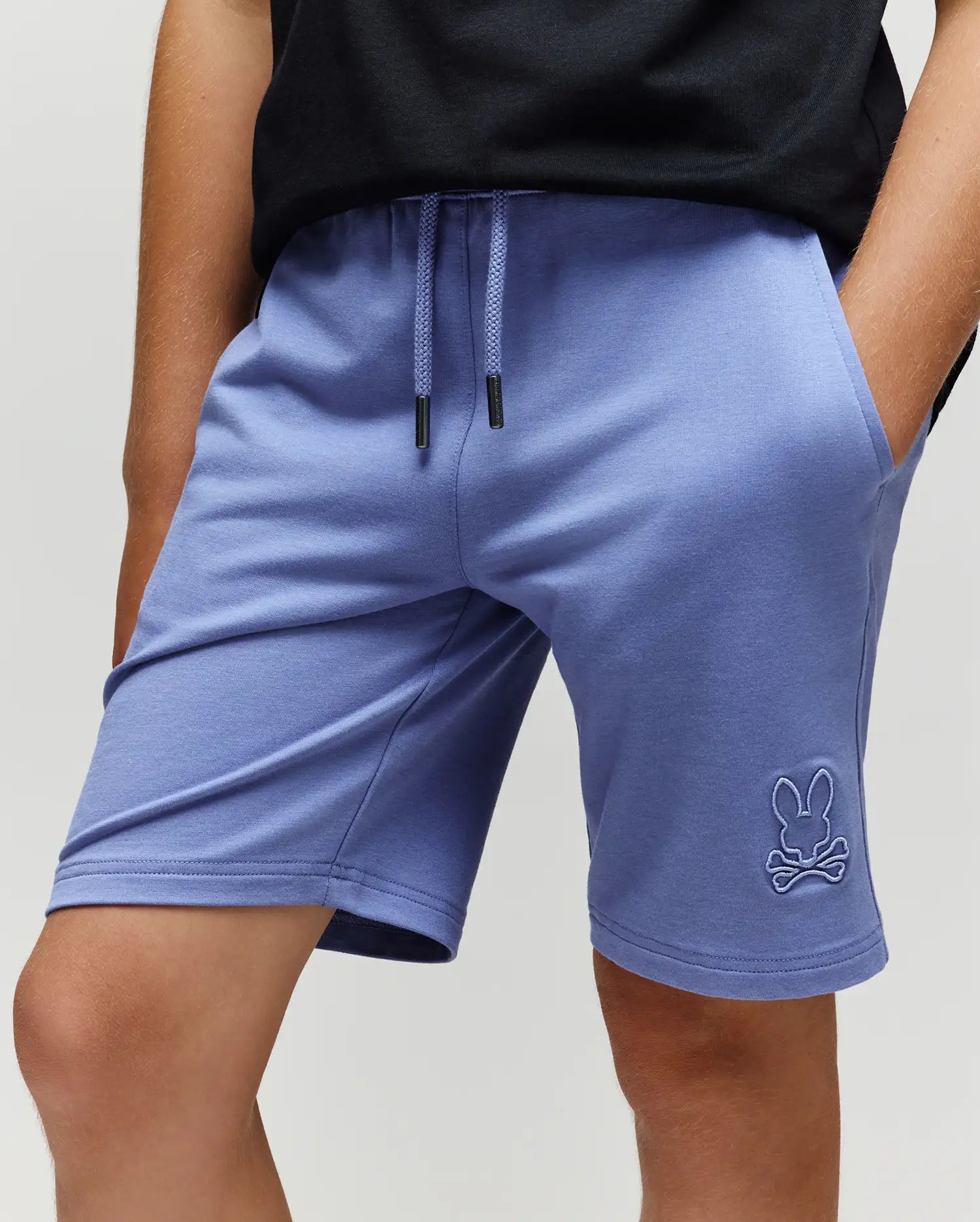 A child wears Psycho Bunny's Kids Levanzo French Terry Sweatshorts (B0R578F200) in light purple, featuring an embroidered Bunny skull logo on the left leg, paired with a black t-shirt and hands in their pockets.