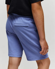 A person is shown from behind waist down, wearing Psycho Bunny KIDS LEVANZO FRENCH TERRY SWEATSHORT - B0R578F200 in light blue with micro French terry fabric, a single back welt pocket, and a black T-shirt; one hand rests by their side.