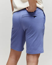 A child is shown from the back wearing Psycho Bunny's KIDS LEVANZO FRENCH TERRY SWEATSHORT - B0R578F200 in light purple with a white shirt. The soft micro French terry shorts have a zippered back pocket. Background is plain and light-colored.
