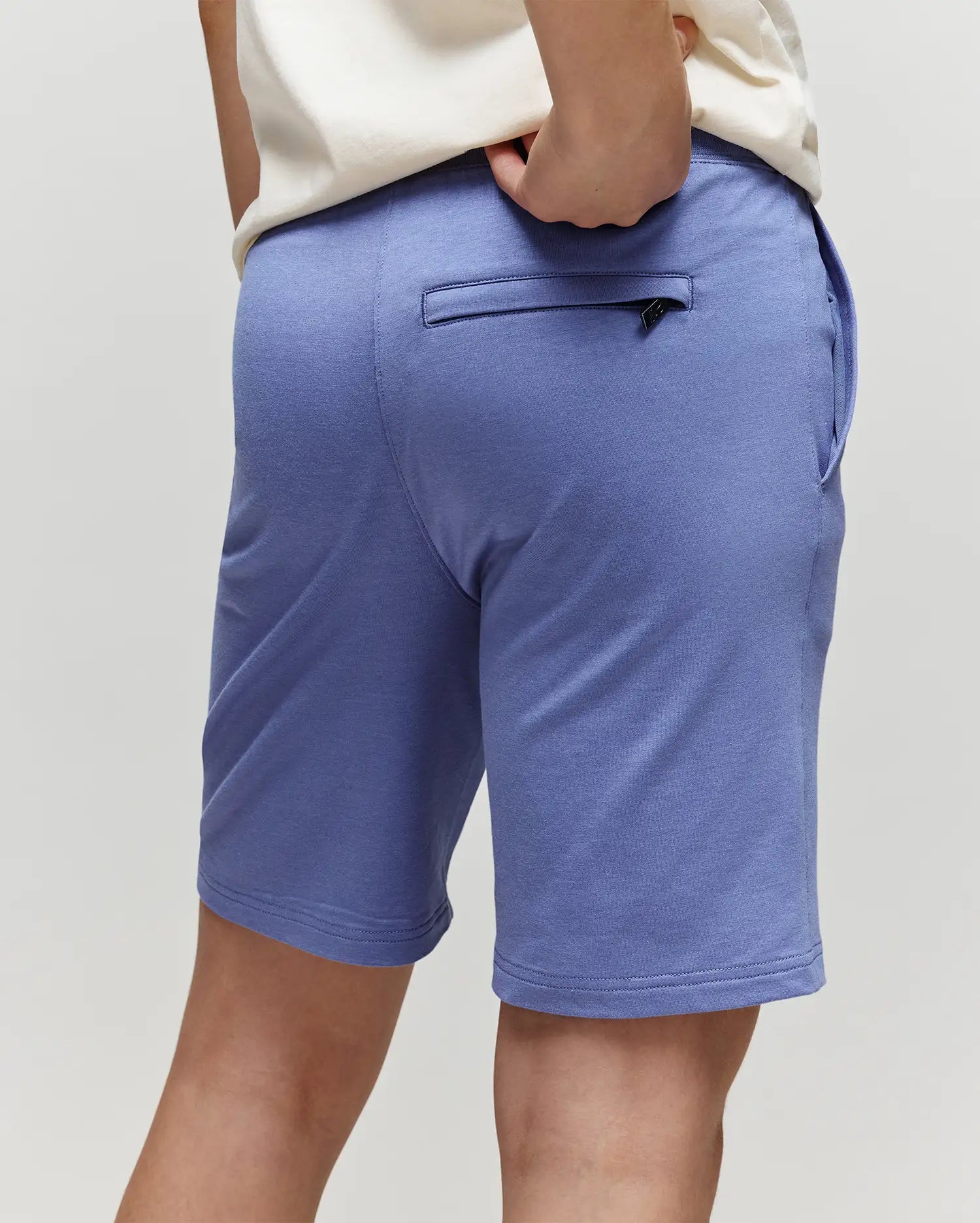 A child is shown from the back wearing Psycho Bunny's KIDS LEVANZO FRENCH TERRY SWEATSHORT - B0R578F200 in light purple with a white shirt. The soft micro French terry shorts have a zippered back pocket. Background is plain and light-colored.