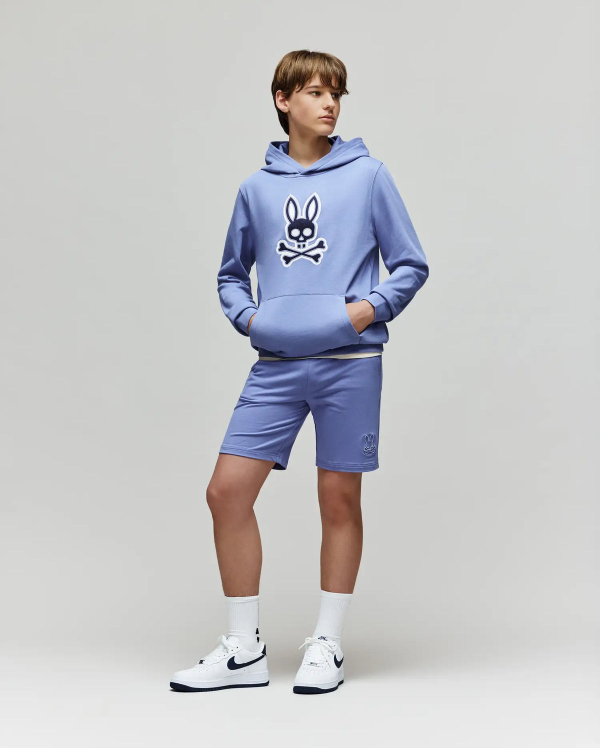 A teenage boy wears Psycho Bunny's KIDS LEVANZO FRENCH TERRY SWEATSHORT (B0R578F200) and a light purple micro French terry hoodie with an embroidered Bunny and crossbones logo, standing with hands in his hoodie pockets.