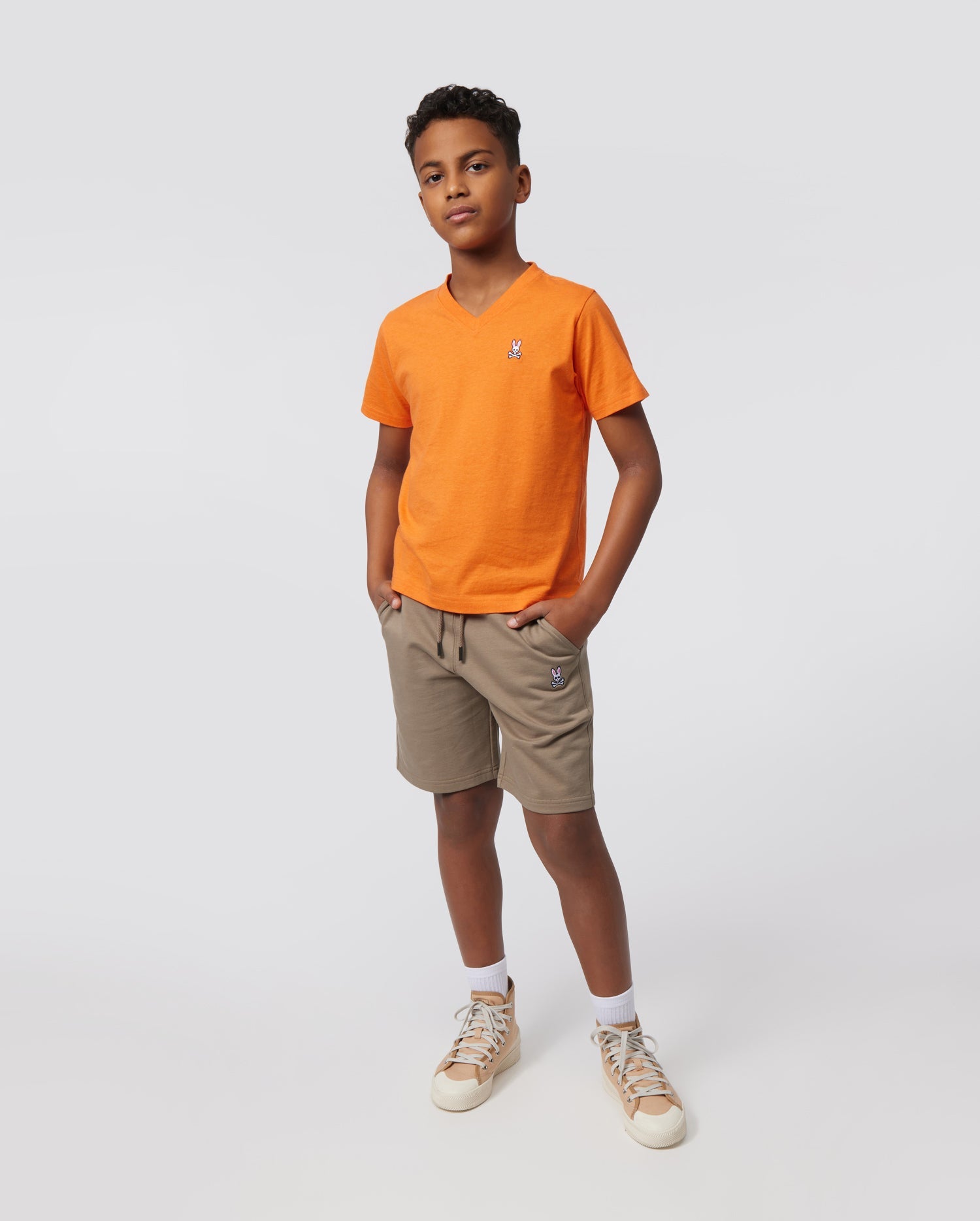 A young boy stands confidently with his hands in the pockets of his beige KIDS FRENCH TERRY SHORTS - B0R829ARFT from Psycho Bunny. He is wearing an orange V-neck T-shirt and white sneakers with high white socks. The background is plain and light gray.
