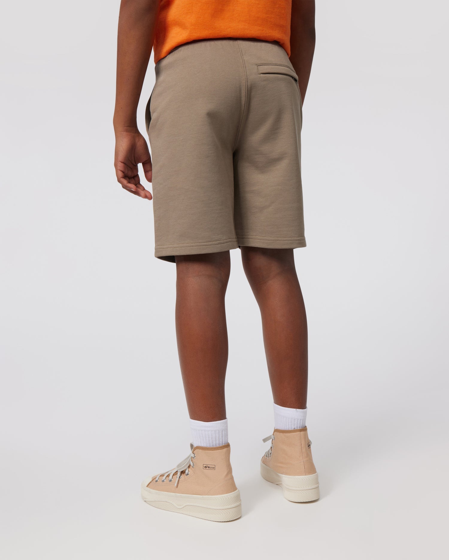 A person wearing a bright orange t-shirt, white socks, and beige high-top sneakers stands with their back to the camera, showcasing the Psycho Bunny KIDS FRENCH TERRY SHORTS - B0R829ARFT. The background is plain white.