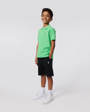 A smiling young boy stands against a plain background, wearing a bright green short-sleeve polo shirt, navy blue KIDS CLASSIC FRENCH TERRY SHORTS - B0R829ARFT by Psycho Bunny with an embroidered Bunny logo, white socks, and white sneakers. He has his hands in his pockets and is looking at the camera.