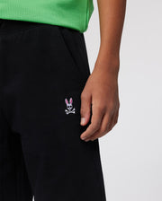 A person wearing a green shirt and black pants. The pants, reminiscent of KIDS CLASSIC FRENCH TERRY SHORTS - B0R829ARFT by Psycho Bunny in their playful design, have an embroidered bunny logo with pink ears and white features positioned near the right pocket. Only the lower torso and hand are visible.
