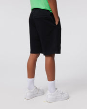 A person wearing a green shirt, white socks, and white sneakers stands with their hands in their pockets. The background is plain and light-colored. The person is shown from the back, focusing on their legs and lower torso. They are also wearing black KIDS CLASSIC FRENCH TERRY SHORTS - B0R829ARFT by Psycho Bunny, featuring an embroidered Bunny logo.