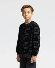 A young boy with curly hair is pictured wearing the Psycho Bunny KIDS ROMEO LIGHTWEIGHT SWEATSHIRT, featuring a bunny-patterned design. He stands facing forward with a neutral expression against a plain, light background.