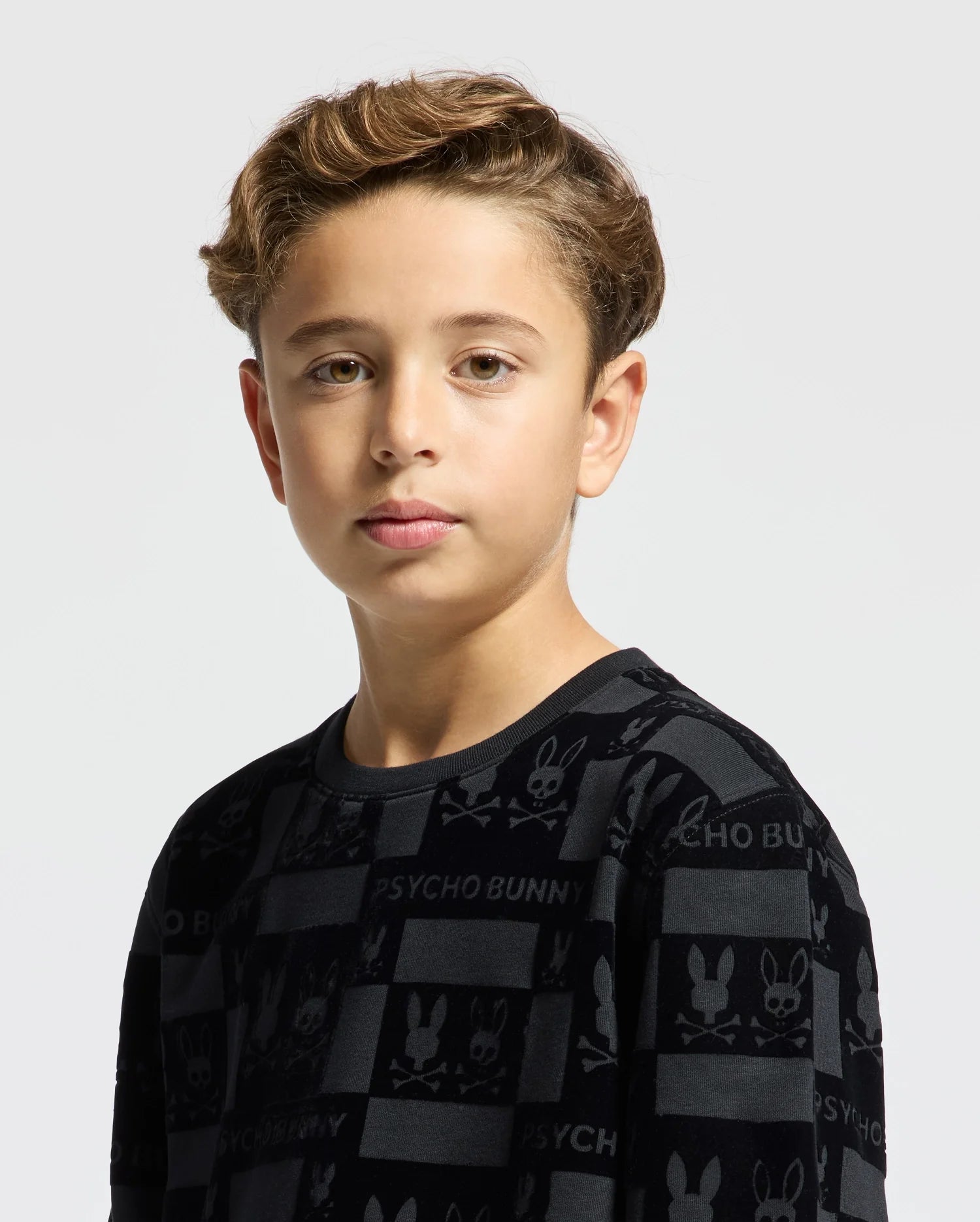 A young person with short brown hair is seen against a plain white background, wearing the KIDS ROMEO LIGHTWEIGHT SWEATSHIRT - B0S129E200 from Psycho Bunny. The sweatshirt features a dark bunny-patterned design with repeated "Psycho Bunny" text and small bunny and skull motifs, adding an edgy touch to the outfit.