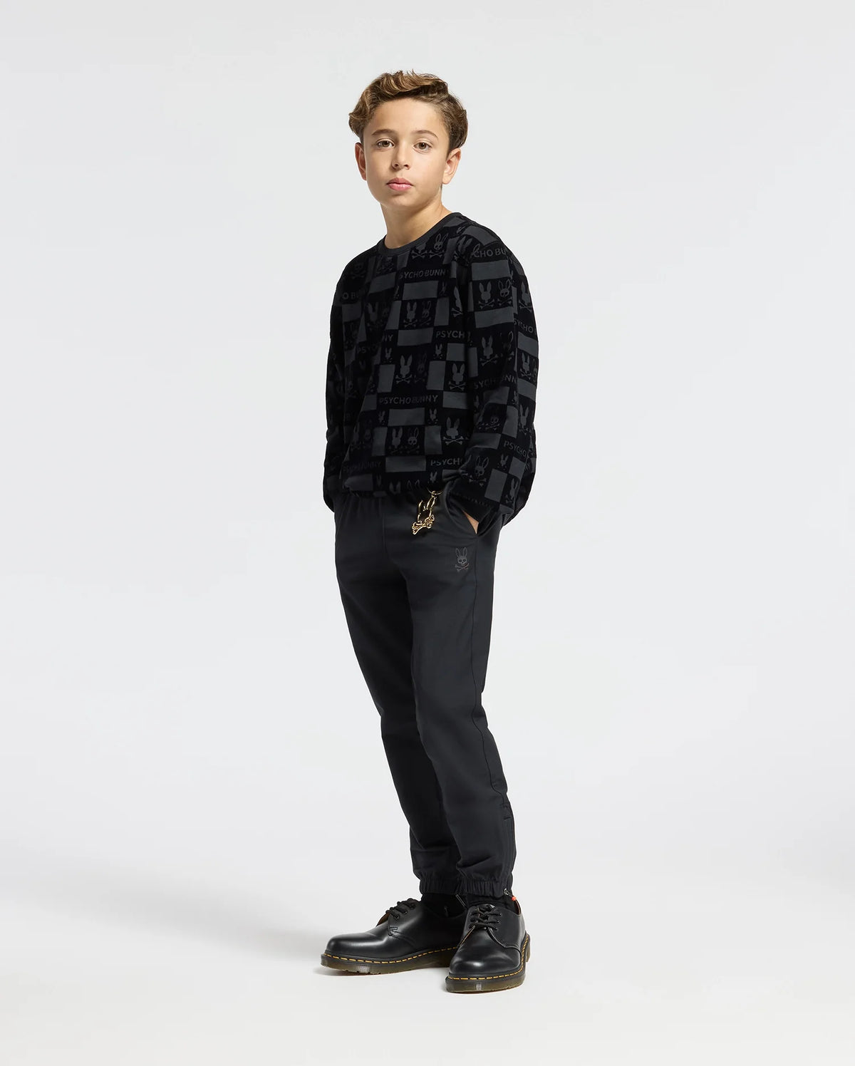 A young person stands against a plain background, wearing the KIDS ROMEO LIGHTWEIGHT SWEATSHIRT by Psycho Bunny, paired with dark pants and black shoes. They have short brown hair and their hands in their pockets.