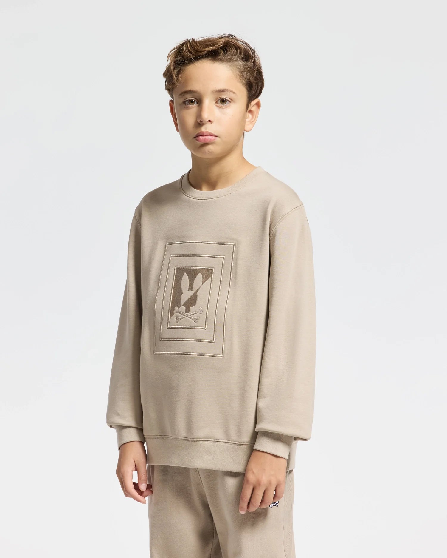 A young person with short hair is wearing a beige Kids Oak City Crew Neck Sweatshirt - B0S274E200 by Psycho Bunny, showcasing a subtle bunny design. The plain, light-colored background enhances the charm of this outfit.
