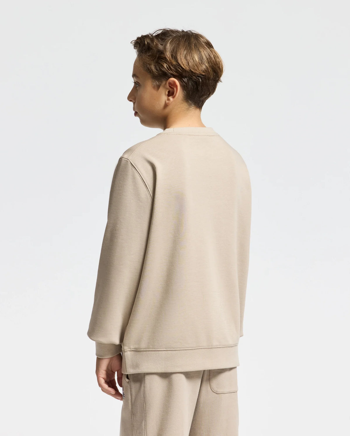 A young person with short brown hair is standing sideways, wearing a Psycho Bunny Kids Oak City Crew Neck Sweatshirt in beige, paired with matching pants against a plain white background.