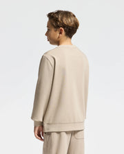 A young person with short brown hair is standing sideways, wearing a Psycho Bunny Kids Oak City Crew Neck Sweatshirt in beige, paired with matching pants against a plain white background.