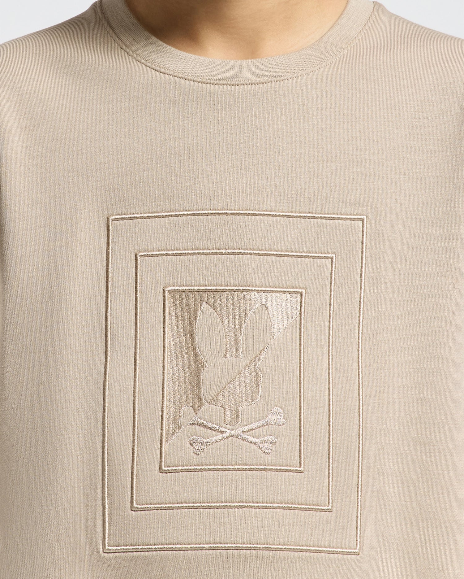 Close-up of the beige Psycho Bunny Kids Oak City Crew Neck Sweatshirt (B0S274E200) showcasing an embroidered design at the center. The motif features a stylized rabbit head above crossed bones, all enclosed within two rectangles, reminiscent of a classic Psycho Bunny style.