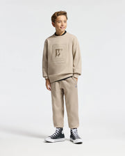 A young boy grins as he stands wearing a KIDS OAK CITY CREW NECK SWEATSHIRT from Psycho Bunny, along with coordinated pants that both highlight a textured design. He's sporting black high-top sneakers with white soles against a plain white backdrop.