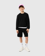 A young boy beams while wearing the Psycho Bunny KIDS HOUSTON FRENCH TERRY CREWNECK - B0S462C200 in black, along with matching black shorts, white sneakers, and a pink cap. He stands against a plain white background with his hands by his sides.