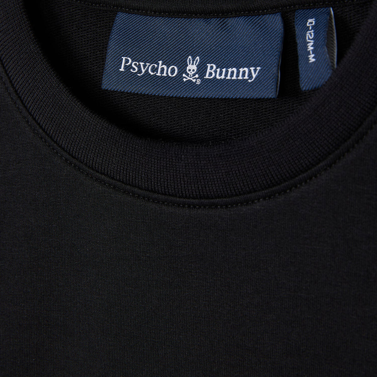 Close-up of a black KIDS HOUSTON FRENCH TERRY CREWNECK (B0S462C200) by Psycho Bunny, showcasing the collar and brand's label inside. Crafted from lightweight Micro French Terry, the label features the signature skull with bunny ears logo, exemplifying the premium cotton fabric quality.
