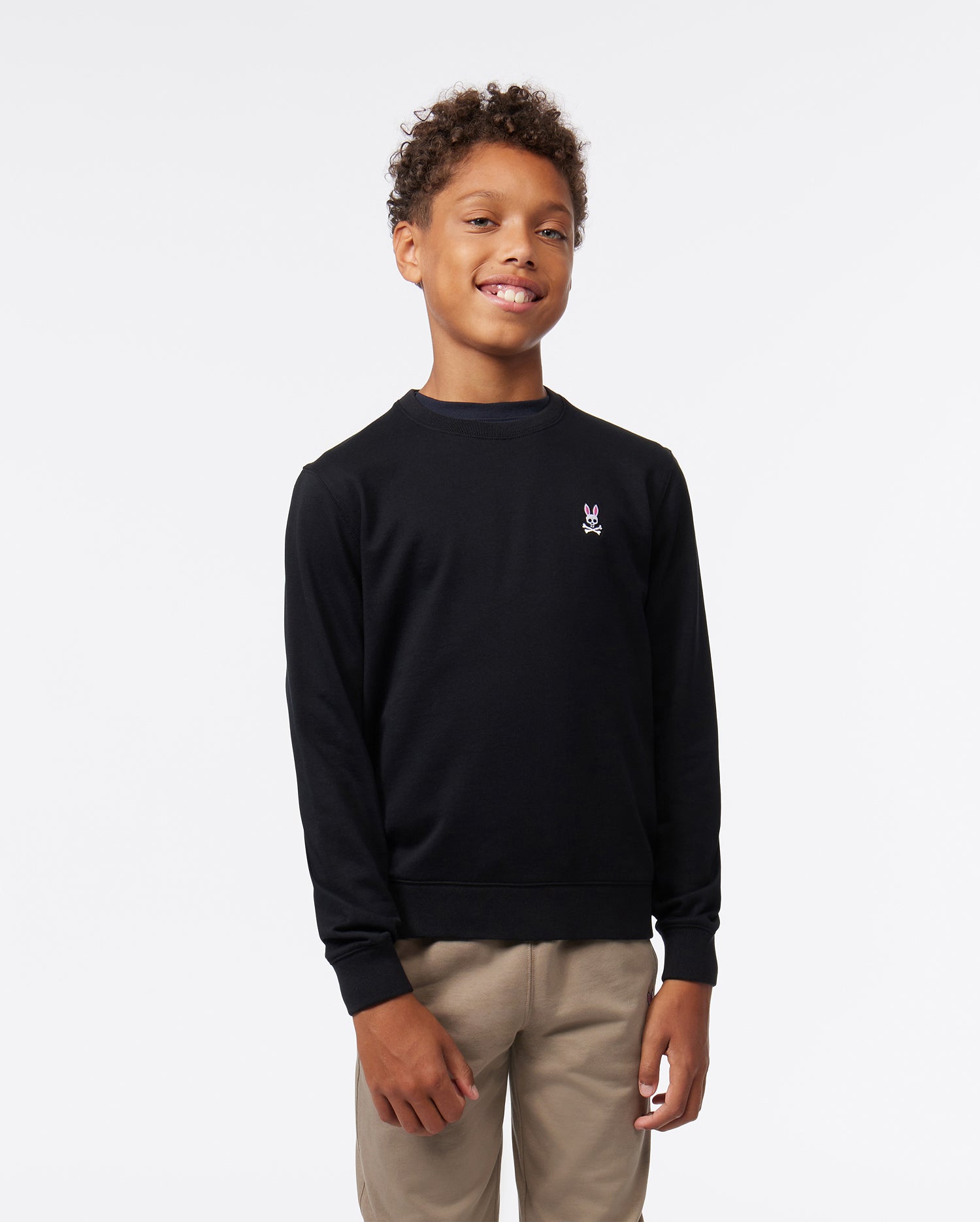 A young boy smiling, wearing a black Psycho Bunny KIDS FRENCH TERRY SWEATSHIRT with an embroidered bunny logo on the left chest and khaki trousers, standing against a white background.