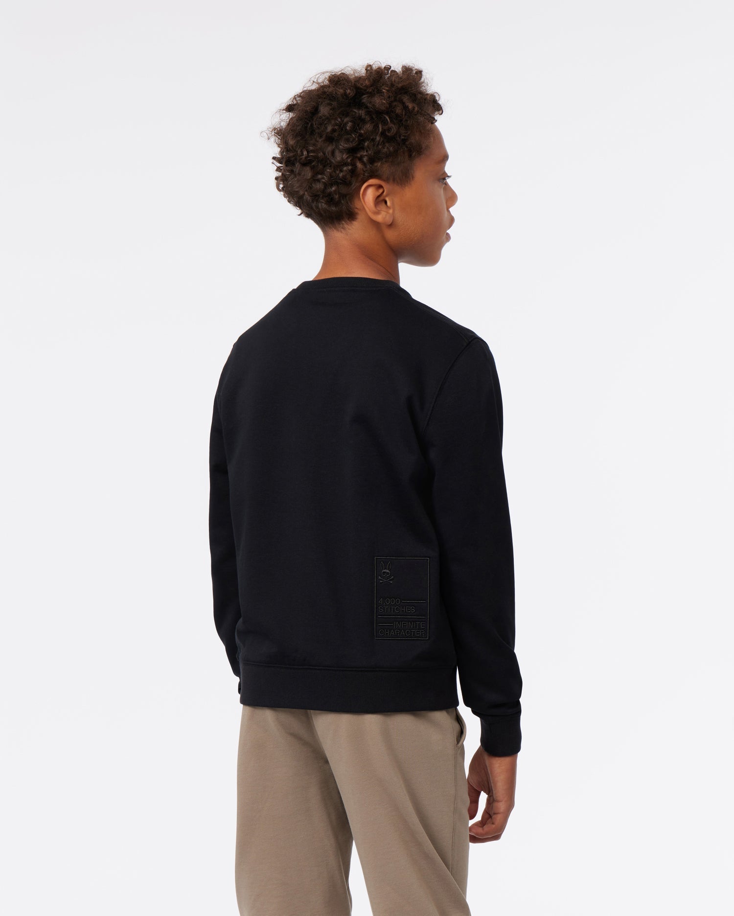 A young boy wearing a dark blue Psycho Bunny KIDS FRENCH TERRY SWEATSHIRT - B0S830ARFT and beige pants, seen from the back, with a focus on the embroidered bunny logo detail on the sweatshirt.