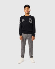 A young individual confidently stands against a plain, light gray background, wearing a KIDS ZACK VARSITY SWEATSHIRT by Psycho Bunny in black with chenille embroidery, paired with gray pants, white sneakers, and a black cap.